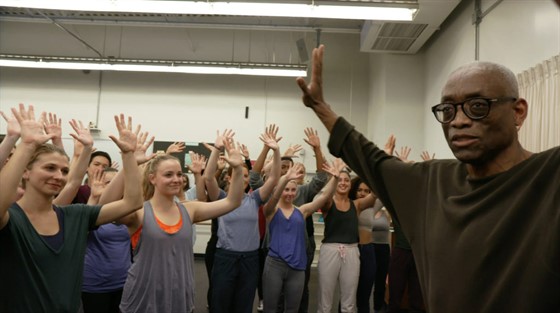 Bill T. Jones tutoring a class of performers, all with hands raised.